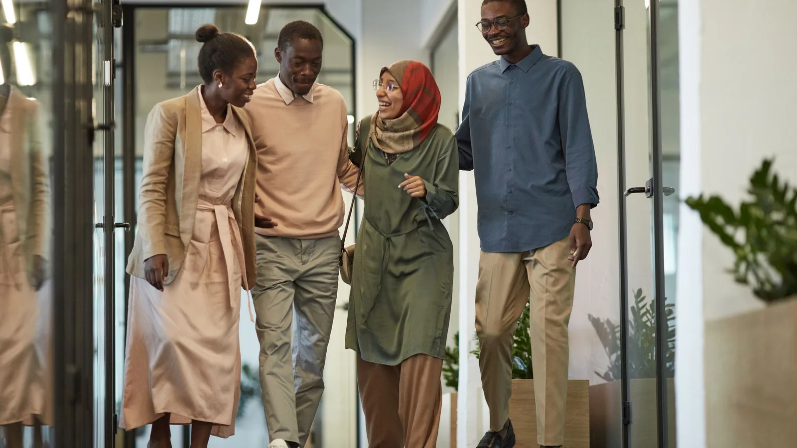Quatre collègues souriants marchant ensemble dans le couloir vitré d'un bâtiment de bureaux
